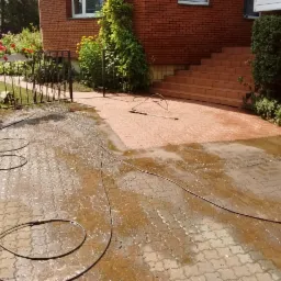 Czerwona kostka brukowa na podjeździe częściowo umyta, widać różnicę w kolorze, wąż ciśnieniowy leży na ziemi, budynek z czerwonej cegły w tle.