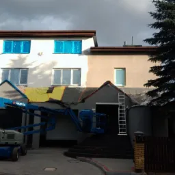 Dwukondygnacyjny dom w trakcie malowania elewacji, z częściowo pomalowaną ścianą w kolorze białym i beżowym, zabezpieczonymi oknami folią, podnośnikiem koszowym PARTA i drabina oparta o ścianę.