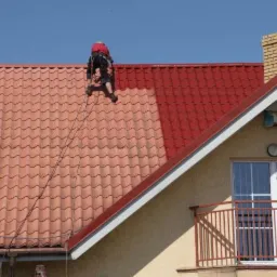 Pracownik w uprzęży zabezpieczającej, maluje dachówkę na dachu domu na kolor czerwony, widoczna połowa dachu pomalowana i połowa w kolorze ceglastym