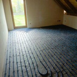 Intal-Tech Systemy grzewcze i sanitarne Maciej Gładysz - Niebieskie rury ogrzewania podłogowego ułożone na siatce w surowym wnętrzu, widok na okno i fragment poddasza.