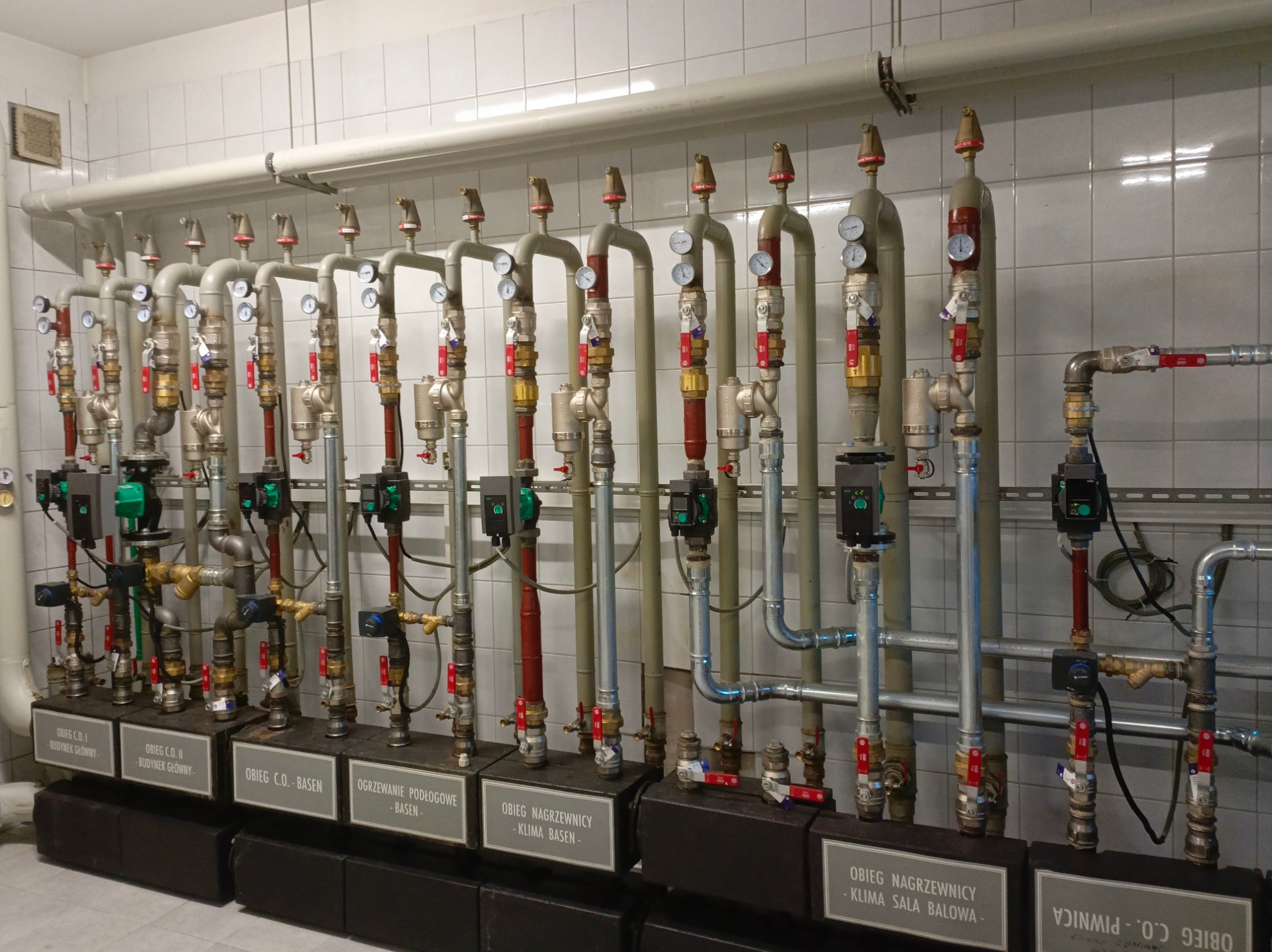 Rozbudowana instalacja grzewcza z licznymi rurami, zaworami, manometrami i pompami obiegowymi w pomieszczeniu wyłożonym białymi płytkami, z etykietami opisującymi poszczególne obiegi grzewcze...