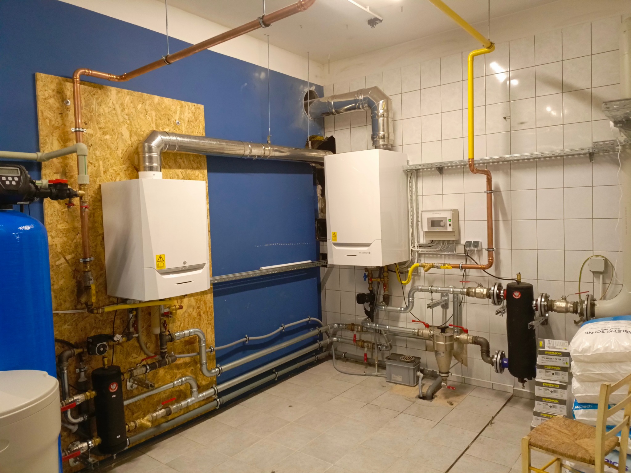 Dwa białe kotły grzewcze wiszące na ścianie z widocznymi rurami miedzianymi i żółtymi, instalacja hydrauliczna w pomieszczeniu z biało-niebieskimi ścianami i ceramiczną podłogą.