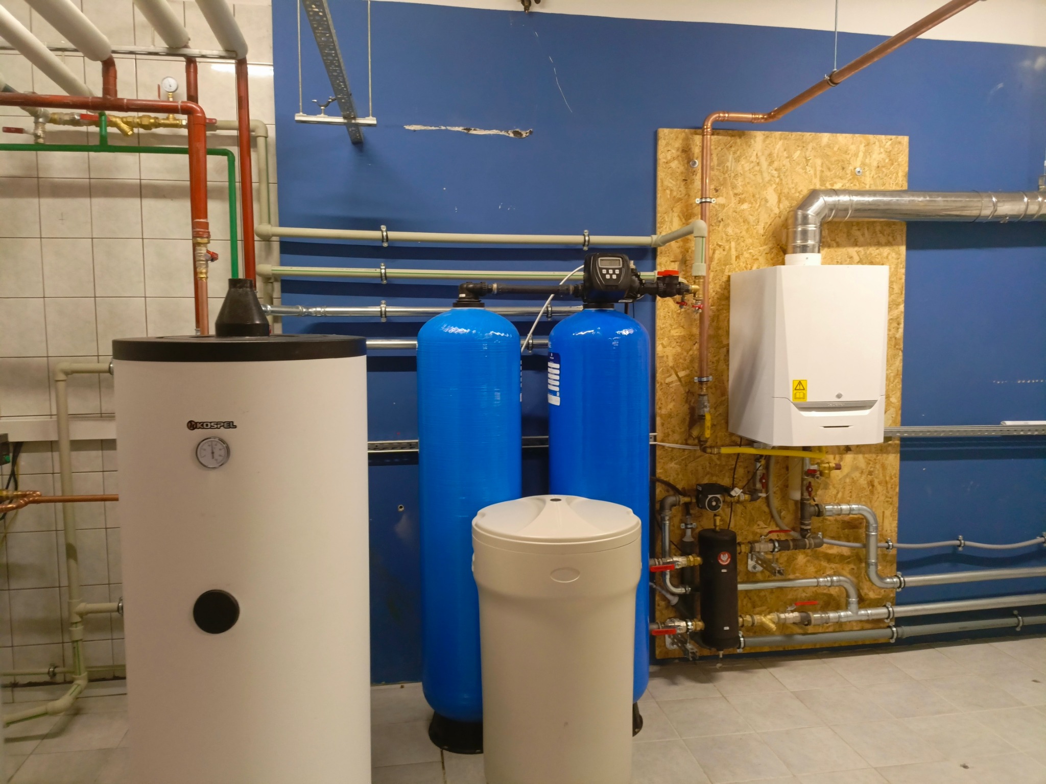 Kotłownia z piecem gazowym, zasobnikiem ciepłej wody, dwoma niebieskimi filtrami i zmiękczaczem wody na tle niebieskiej ściany i płytek.
