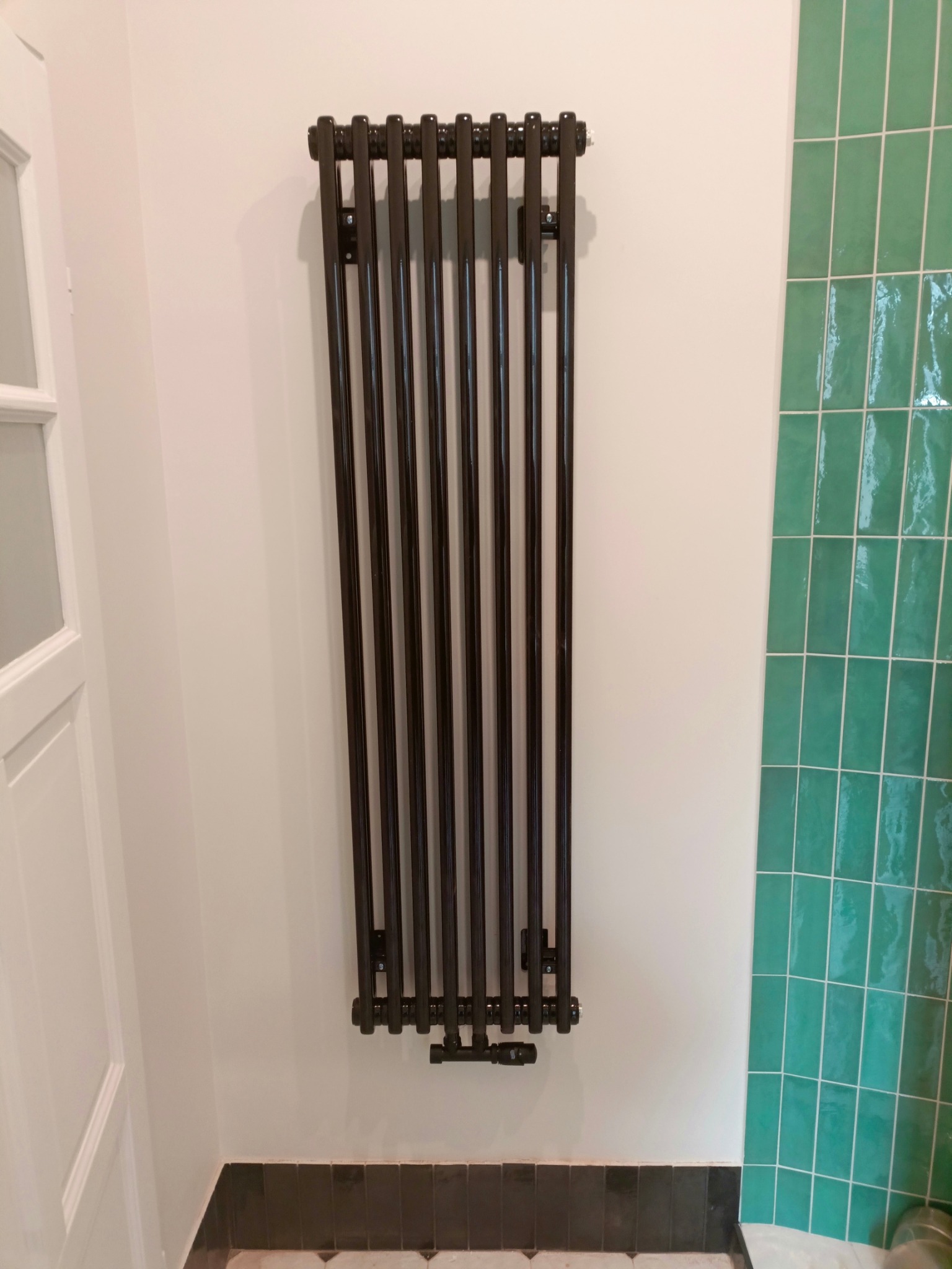 Czarny, pionowy grzejnik żeberkowy zamontowany na białej ścianie, widoczne zielone płytki w tle i fragment białych drzwi po lewej.