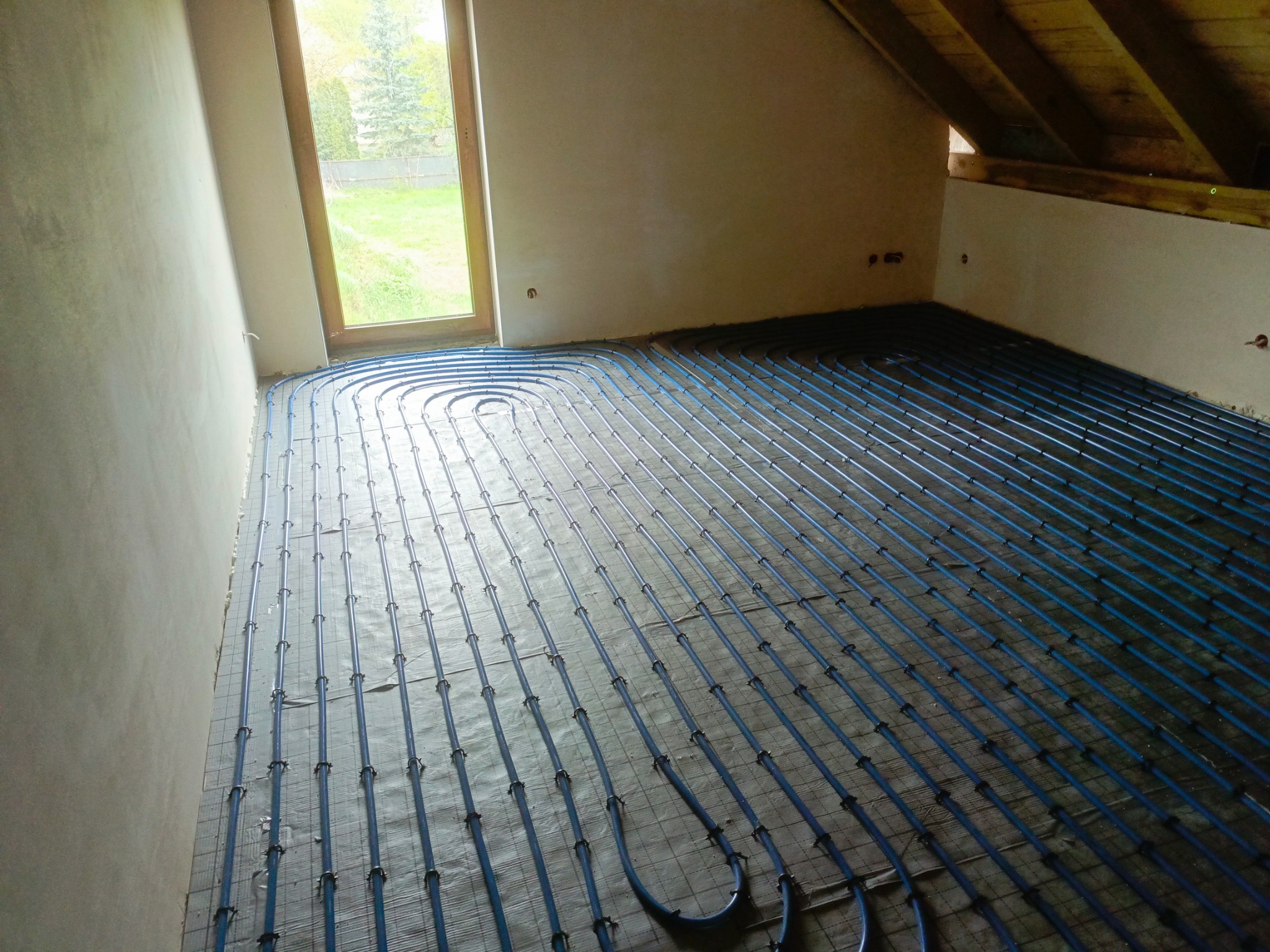 Niebieskie rury ogrzewania podłogowego ułożone na siatce w surowym wnętrzu, widok na okno i fragment poddasza.