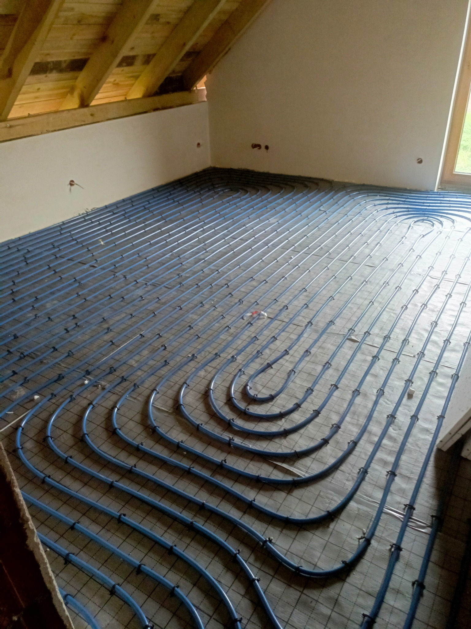 Niebieskie rury ogrzewania podłogowego ułożone w pętle na czarnej siatce w przestrzeni poddasza ze skośnym sufitem i drewnianą konstrukcją dachu.