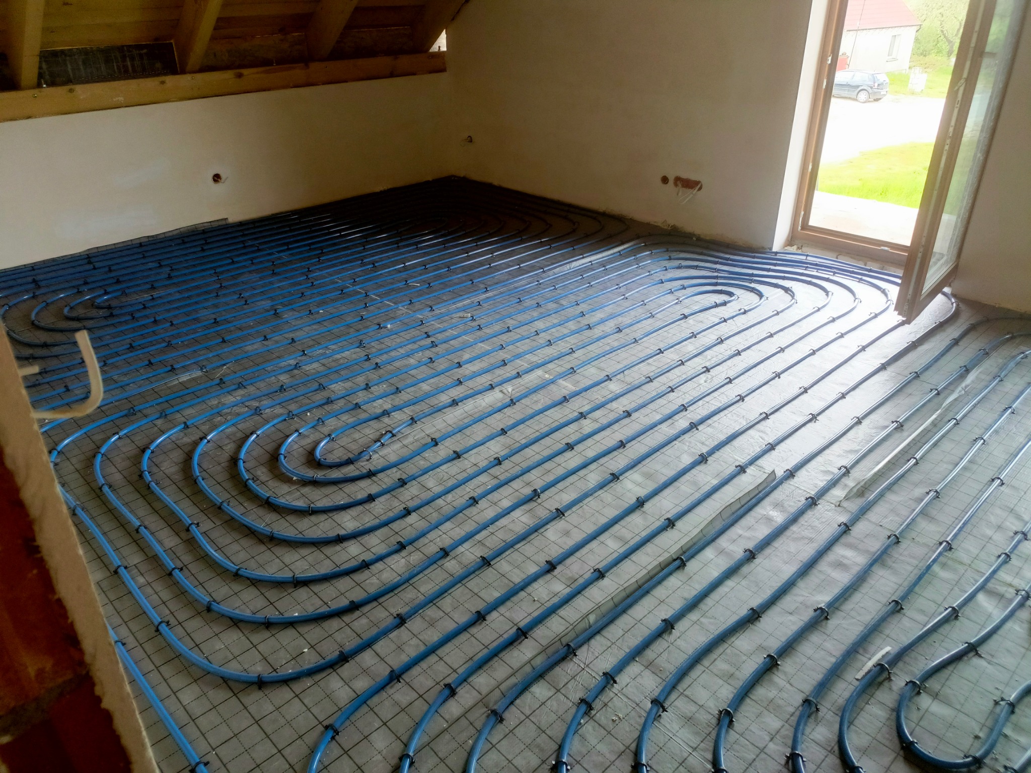 Niebieskie rury ogrzewania podłogowego ułożone spiralnie na siatce w surowym wnętrzu, widoczne fragmenty poddasza i okno z widokiem na zewnątrz.