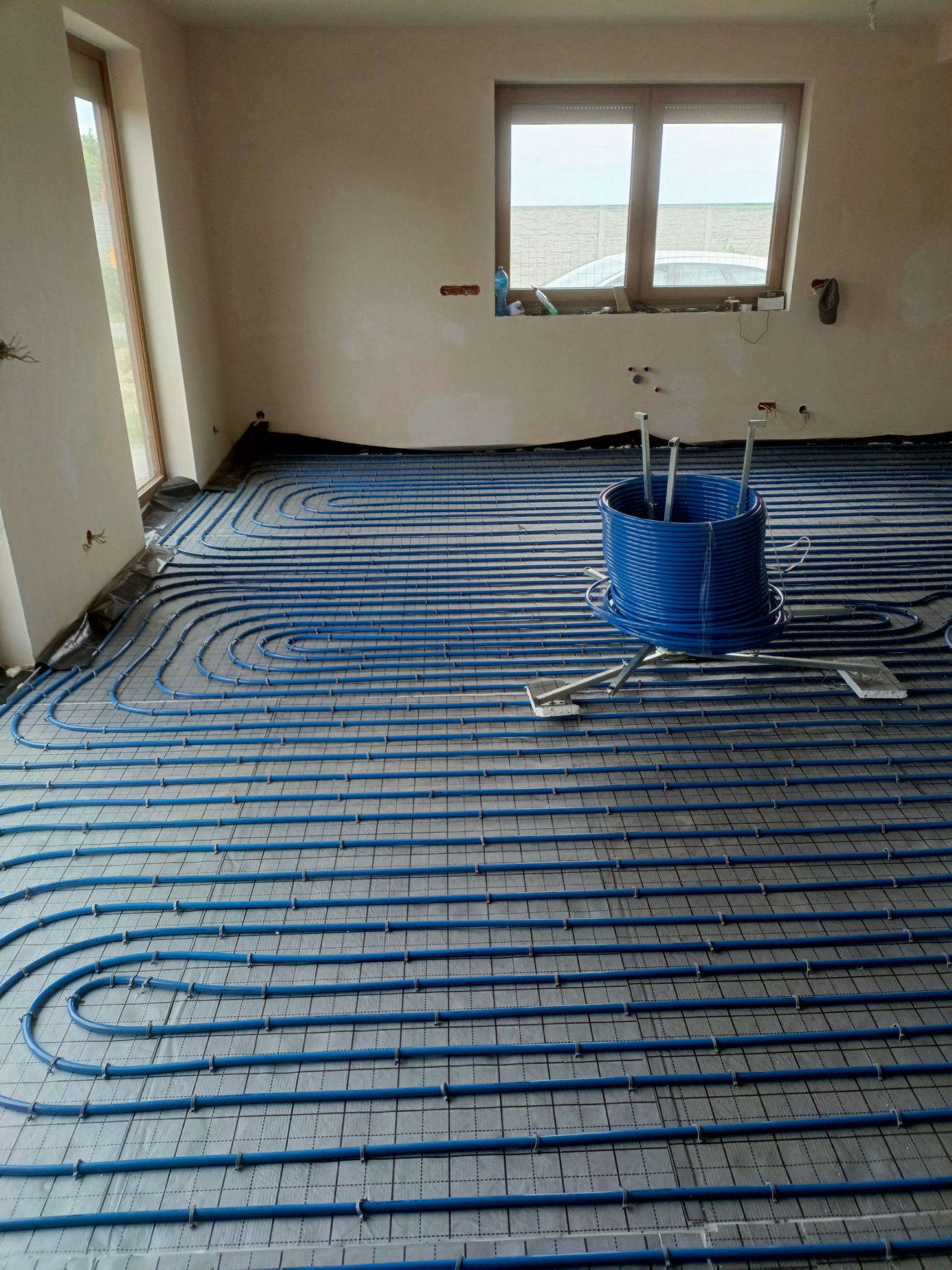 Niebieskie rury ogrzewania podłogowego ułożone na szarej macie w surowym wnętrzu, zwój rur na metalowym stojaku w tle, widok na okno z parapetem zagraconym przedmiotami.