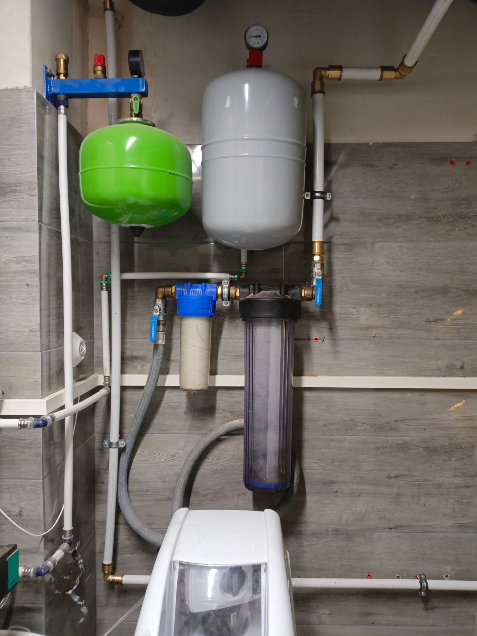 Instalacja wodna z zielonym i szarym zbiornikiem wyrównawczym, filtrami wody i białym zmiękczaczem wody na tle ściany imitującej drewno.