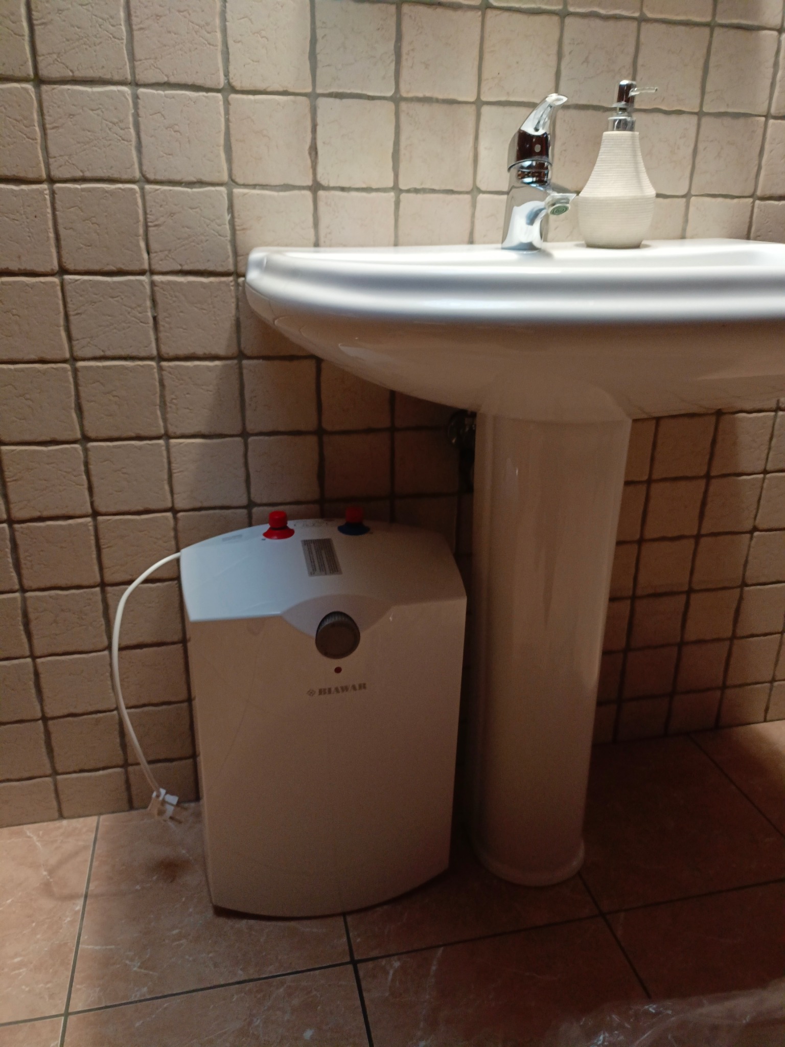 Biały, kompaktowy podgrzewacz wody marki Biawar z czerwono-niebieskimi zaworami, umieszczony obok umywalki na postumencie w łazience z beżowymi płytkami.