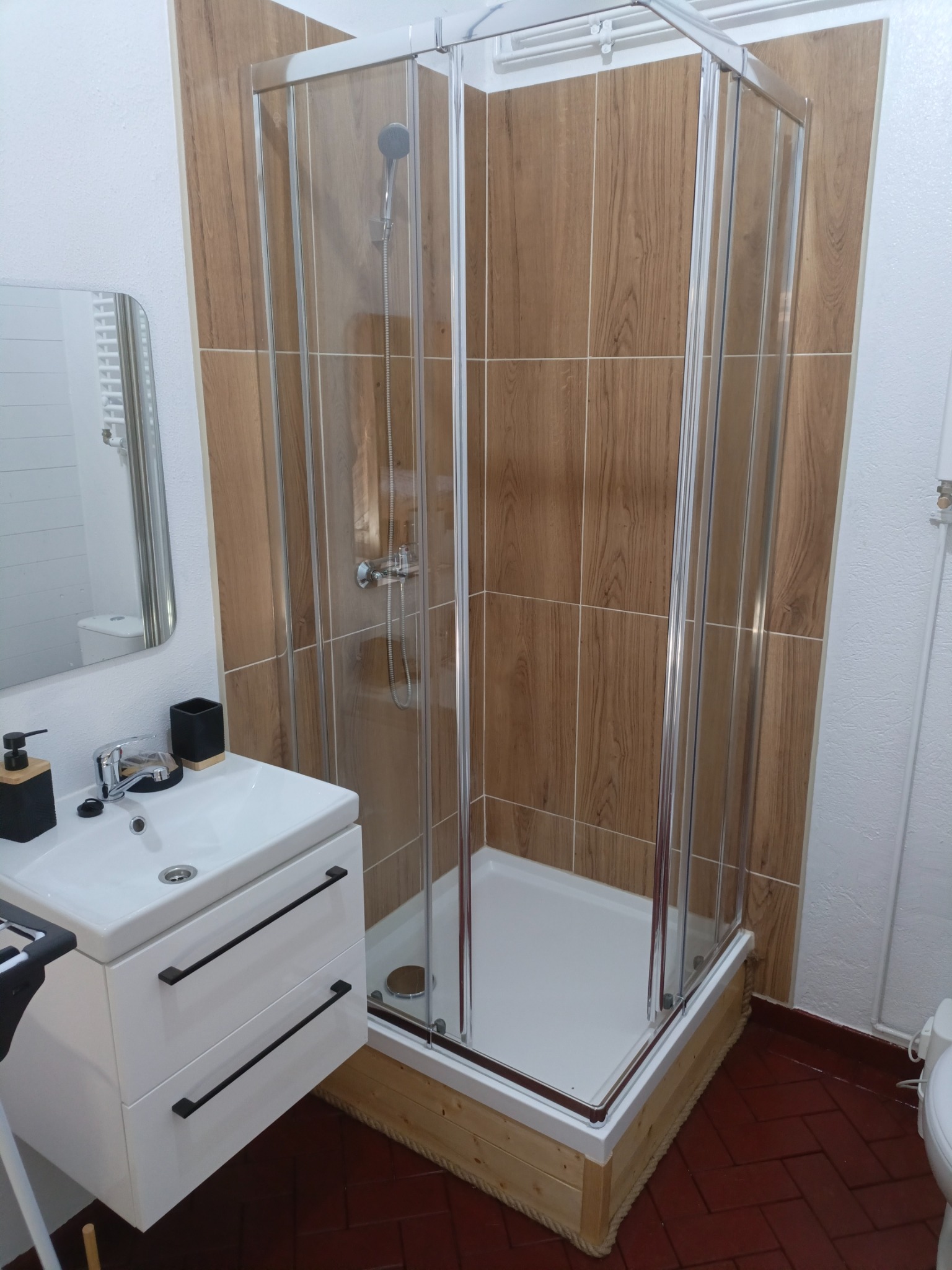 Nowoczesna kabina prysznicowa z drewnopodobnymi płytkami, biała umywalka z czarnymi akcentami i lustro w tle. Ciemna, ceglana podłoga.