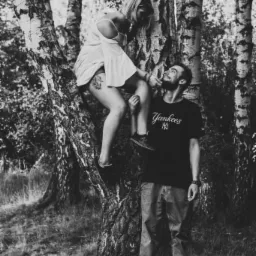 Czarno-białe zdjęcie pary w lesie: kobieta siedzi na gałęzi brzozy, mężczyzna podtrzymuje jej stopę, oboje patrzą na siebie z uśmiechem.