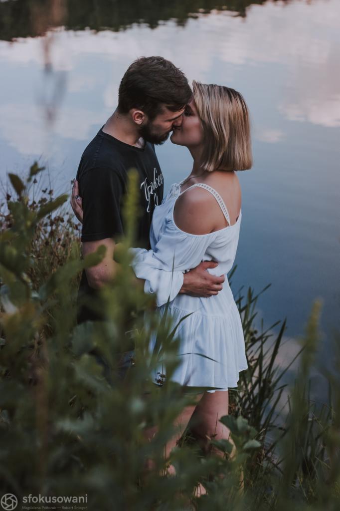 Zakochana para całuje się na tle jeziora, widoczna przez zarośla, kobieta w białej sukience z odkrytymi ramionami, mężczyzna w czarnym t-shircie.