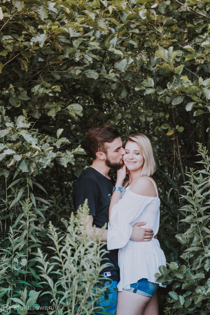 Zakochana para w plenerze: mężczyzna całuje kobietę w policzek na tle zieleni, kobieta w białej bluzce i jeansowych spodenkach, mężczyzna w czarnym t-shircie.