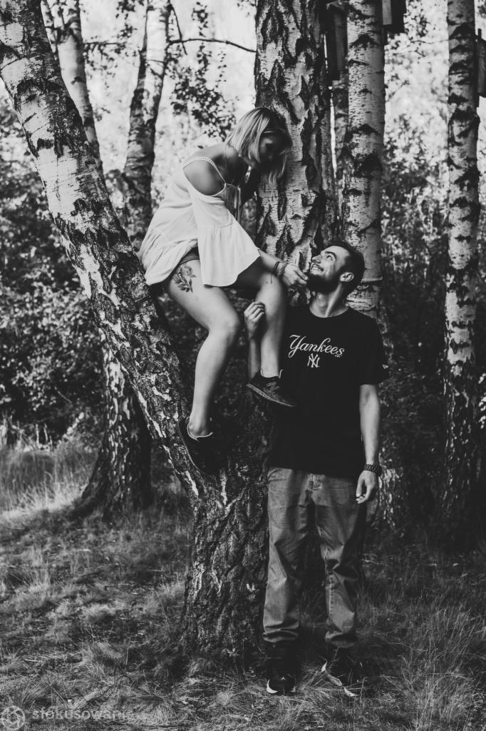 Czarno-białe zdjęcie pary w lesie: kobieta siedzi na gałęzi brzozy, mężczyzna podtrzymuje jej stopę, oboje patrzą na siebie z uśmiechem.