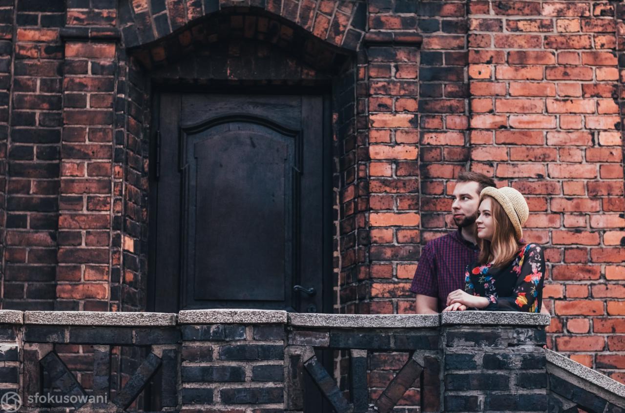 Para zakochanych stoi na ceglanym balkonie przed ciemnymi drzwiami, ona w kwiecistej sukience i słomkowym kapeluszu, on w koszuli w kratę, oboje patrzą w dal.