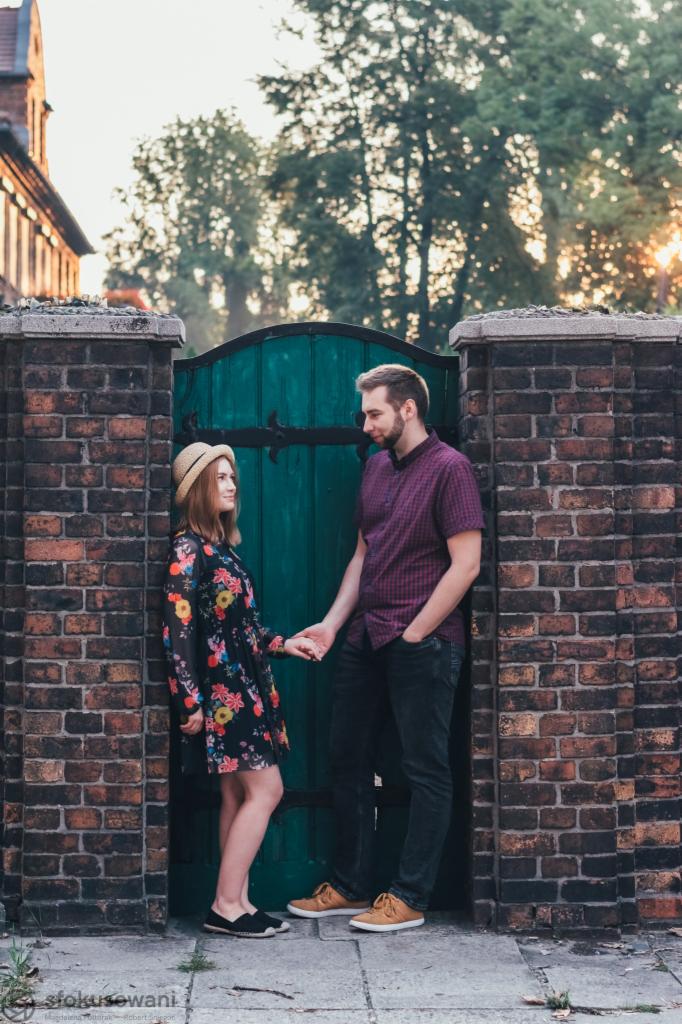 Zakochana para trzymająca się za ręce na tle ceglanego muru i zielonej bramy w Mysłowicach, ona w kwiecistej sukience i kapeluszu, on w koszuli w kratę, ciepłe światło zachodzącego słońca.