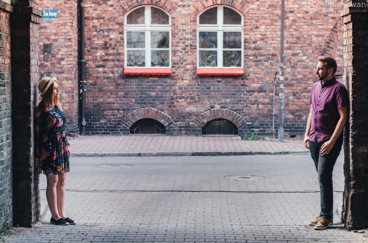 Para stoi po przeciwnych stronach uliczki z ceglanymi budynkami w tle, ona w kwiecistej sukience i kapeluszu, on w koszuli w kratę i jeansach.