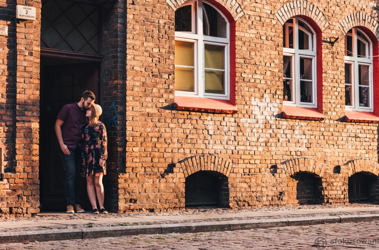 Para zakochanych stoi przy wejściu do ceglanego budynku w promieniach zachodzącego słońca, tworząc intymną atmosferę.