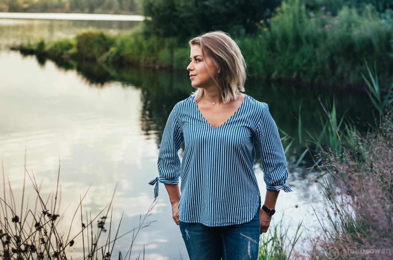 Kobieta w bluzce w paski i jeansach, stojąca nad brzegiem jeziora, patrzy w dal, z widocznym odblaskiem nieba w wodzie i zielenią w tle.