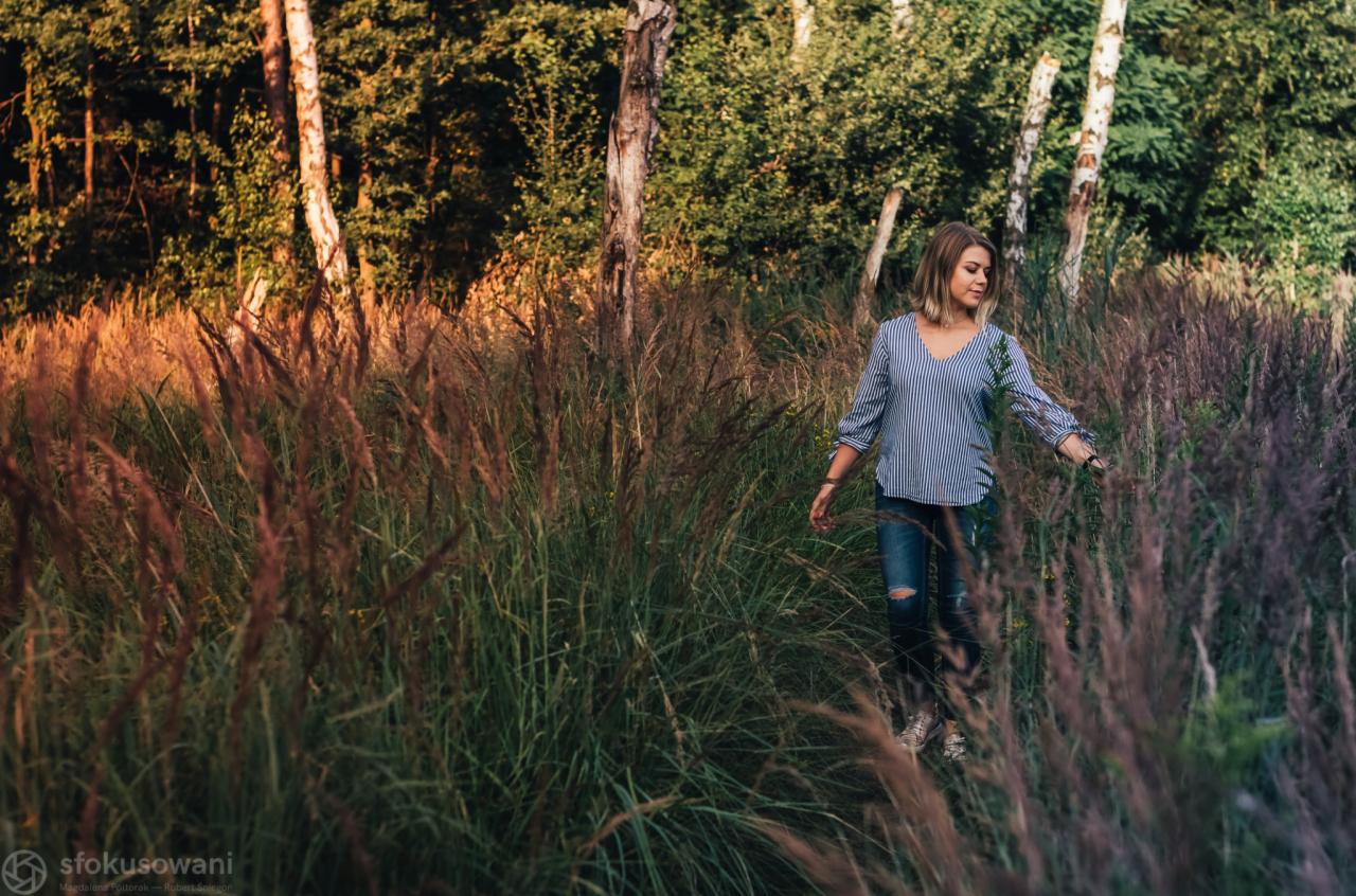Kobieta w bluzce w paski i podartych jeansach idzie przez pole wysokiej trawy w słoneczny dzień, z drzewami brzozowymi w tle.