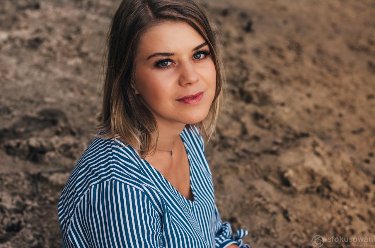 Portret młodej kobiety w bluzce w niebieskie paski, z delikatnym uśmiechem i spojrzeniem w obiektyw, na tle piaszczystej plaży.