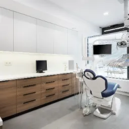 Nowoczesny gabinet stomatologiczny z drewnianymi szafkami, białymi blatami, fotelem dentystycznym z niebieskim obiciem i sprzętem medycznym pod oknem z żaluzjami.