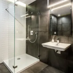 Nowoczesna łazienka w Poznaniu z kabiną prysznicową typu walk-in z białymi, geometrycznymi płytkami, grafitowymi płytkami na ścianach i podłodze oraz podwieszaną umywalką z lustrem podświetlonym...