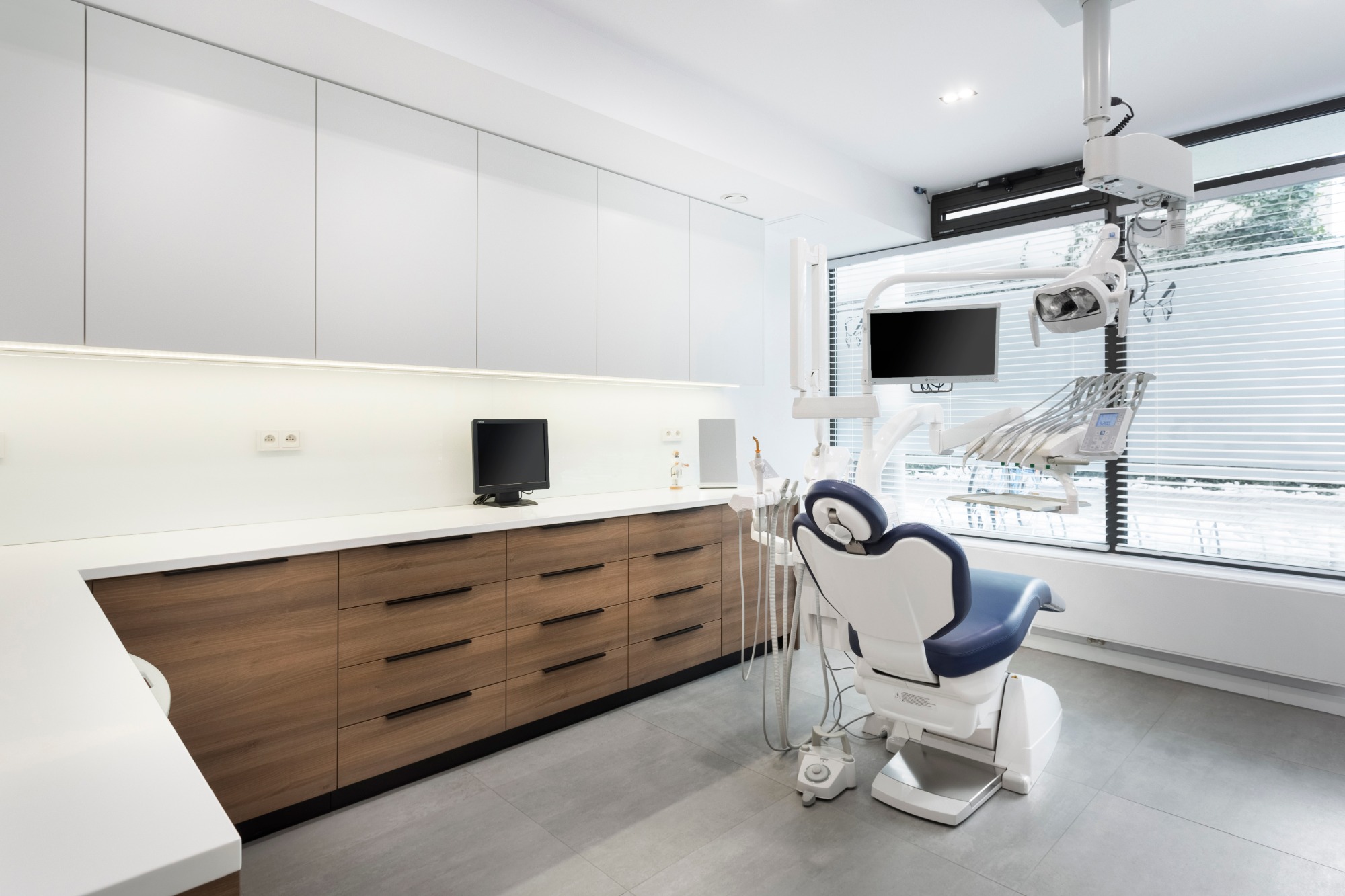 Nowoczesny gabinet stomatologiczny z drewnianymi szafkami, białymi blatami, fotelem dentystycznym z niebieskim obiciem i sprzętem medycznym pod oknem z żaluzjami.