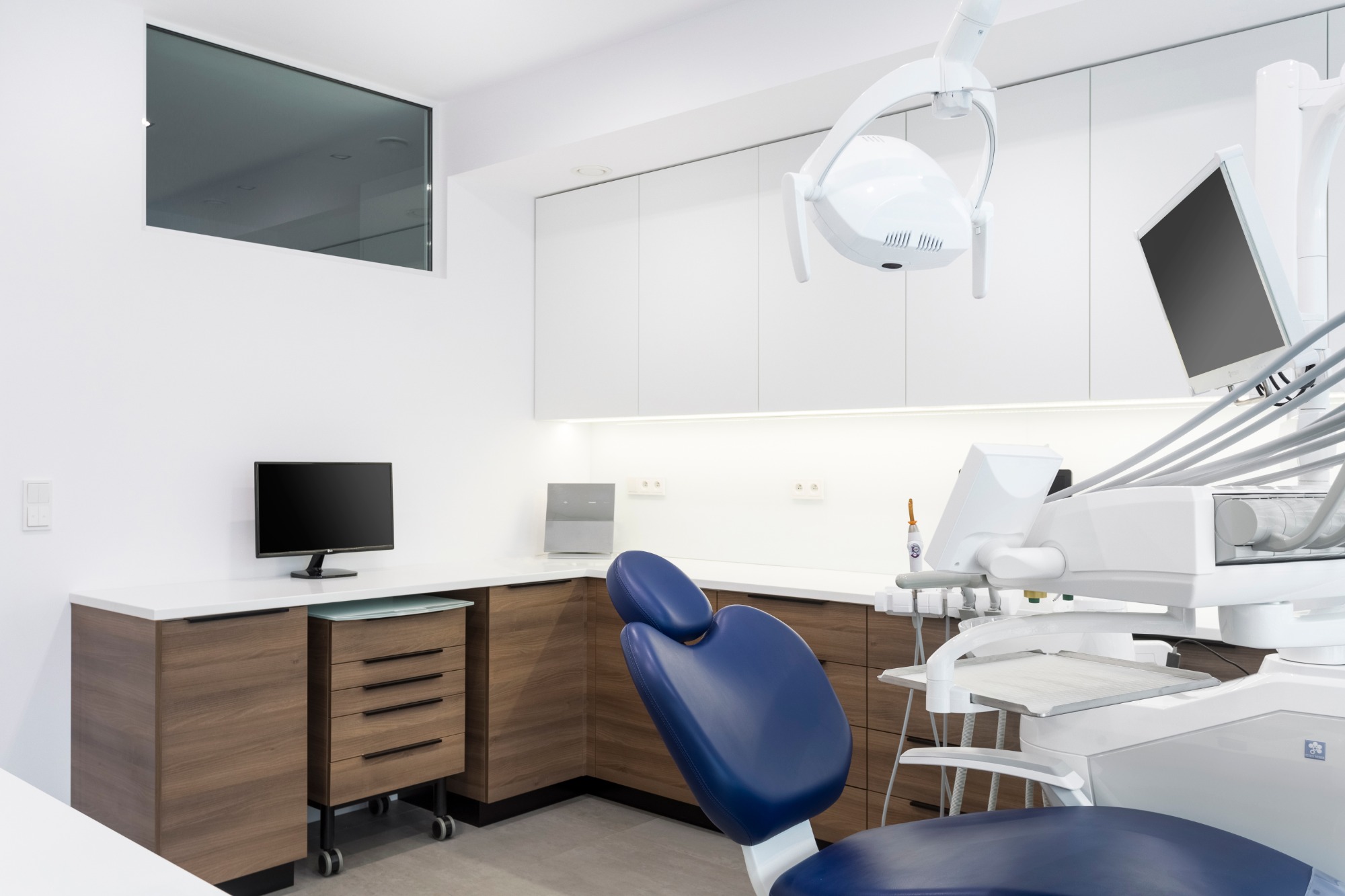 Wnętrze gabinetu stomatologicznego z niebieskim fotelem dentystycznym, białym sprzętem medycznym, drewnianymi szafkami i białymi ścianami, oświetlone lampą LED.