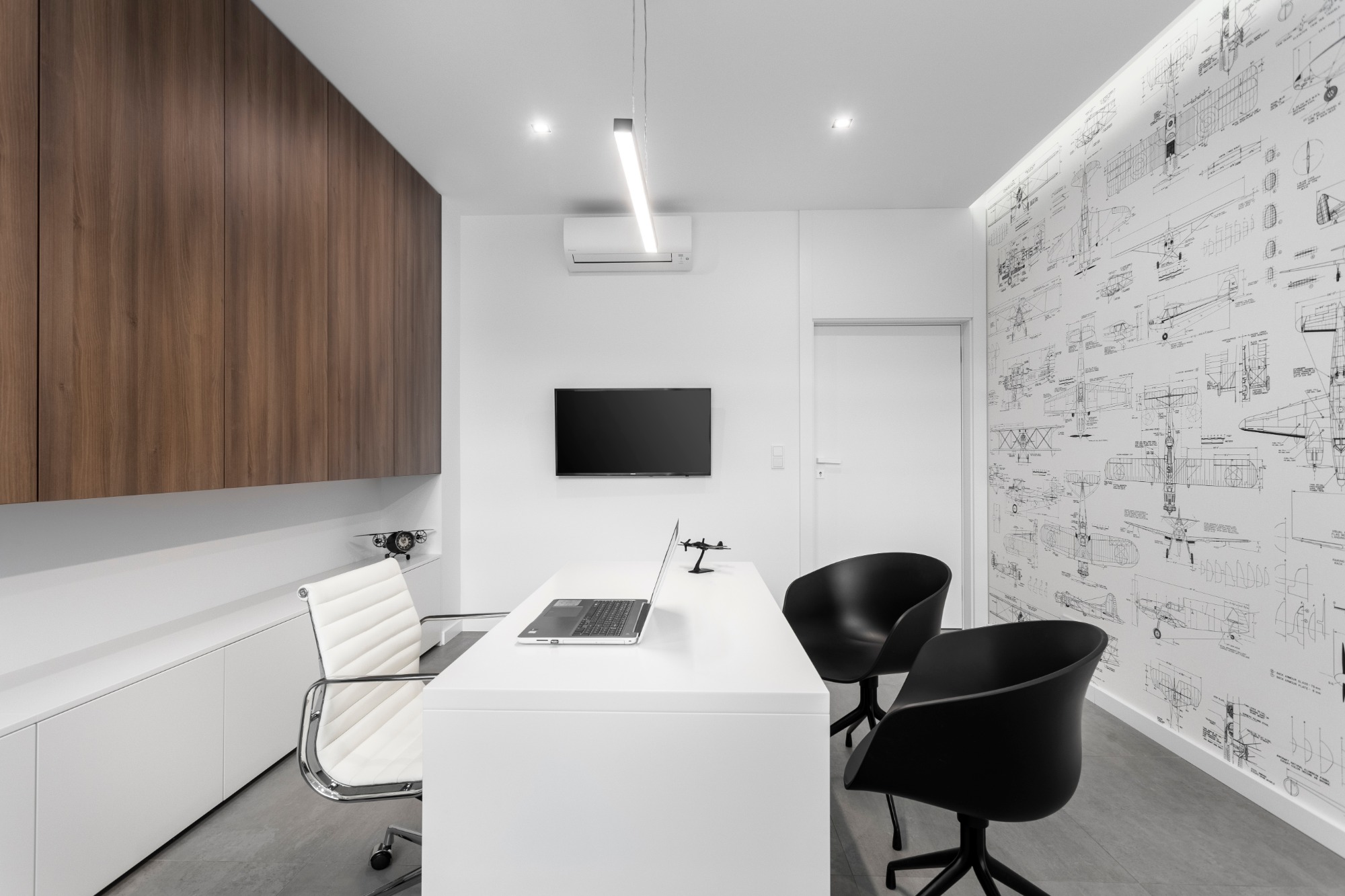 Nowoczesne biuro z białym biurkiem, laptopem, czarnymi krzesłami i ścianą z rysunkami technicznymi samolotów, drewniana zabudowa meblowa na ścianie po lewej stronie.
