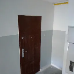 Wnętrze klatki schodowej z brązowymi drzwiami wejściowymi i pomalowanymi na biało-szaro ścianami, widoczny fragment żółtej rury pod sufitem.