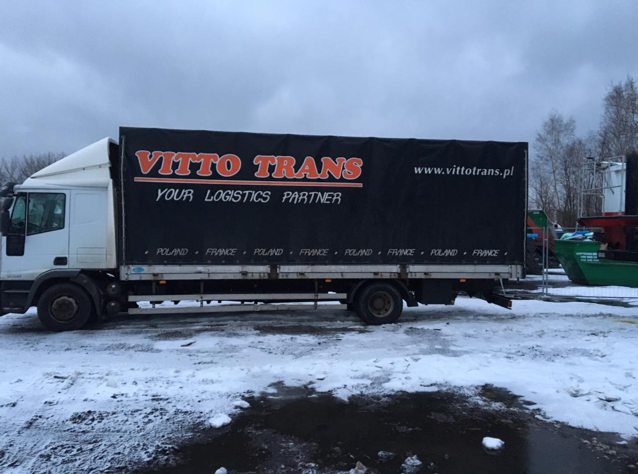 Ciężarówka z czarną plandeką z napisem VITTO TRANS i adresem strony internetowej na tle zimowego krajobrazu ze śniegiem i kałużami.