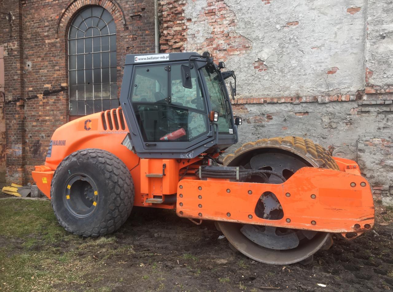 Pomarańczowy walec drogowy HAMM stoi na błotnistym terenie przed starą, ceglaną ścianą budynku z dużym oknem łukowym.