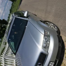 Srebrny samochód osobowy marki Audi, zaparkowany na poboczu asfaltowej drogi, widok z przodu i z góry, w tle białe ogrodzenie i zieleń drzew.