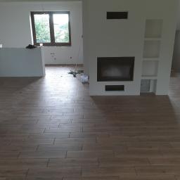 ADIBAU - Salon w trakcie wykańczania: ułożona podłoga z brązowych płytek imitujących drewno, ściany w trakcie malowania, widoczny kominek z wnękami i okno.