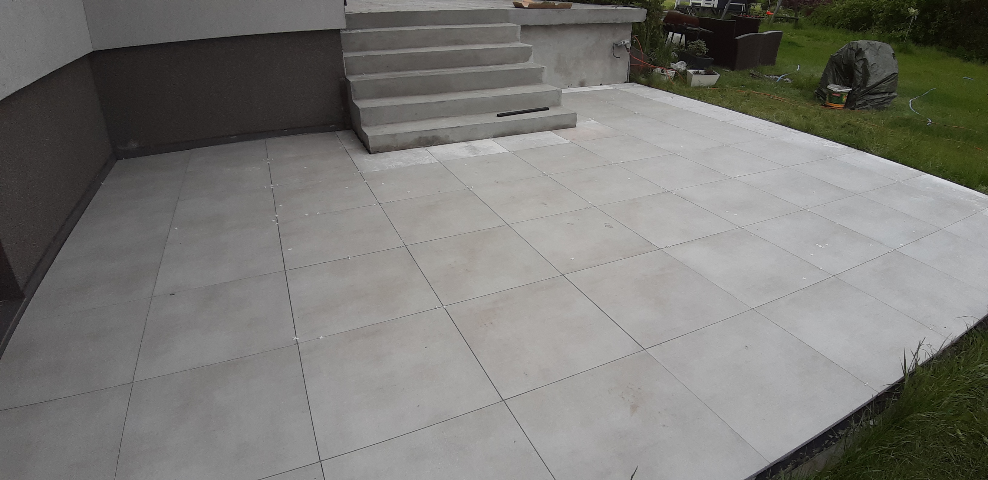 Ułożona jasnoszara płytka gresowa na tarasie zewnętrznym z betonowymi schodami, widoczne krzyżyki dystansowe, na trawniku wiadro i przykryta folią puszka.