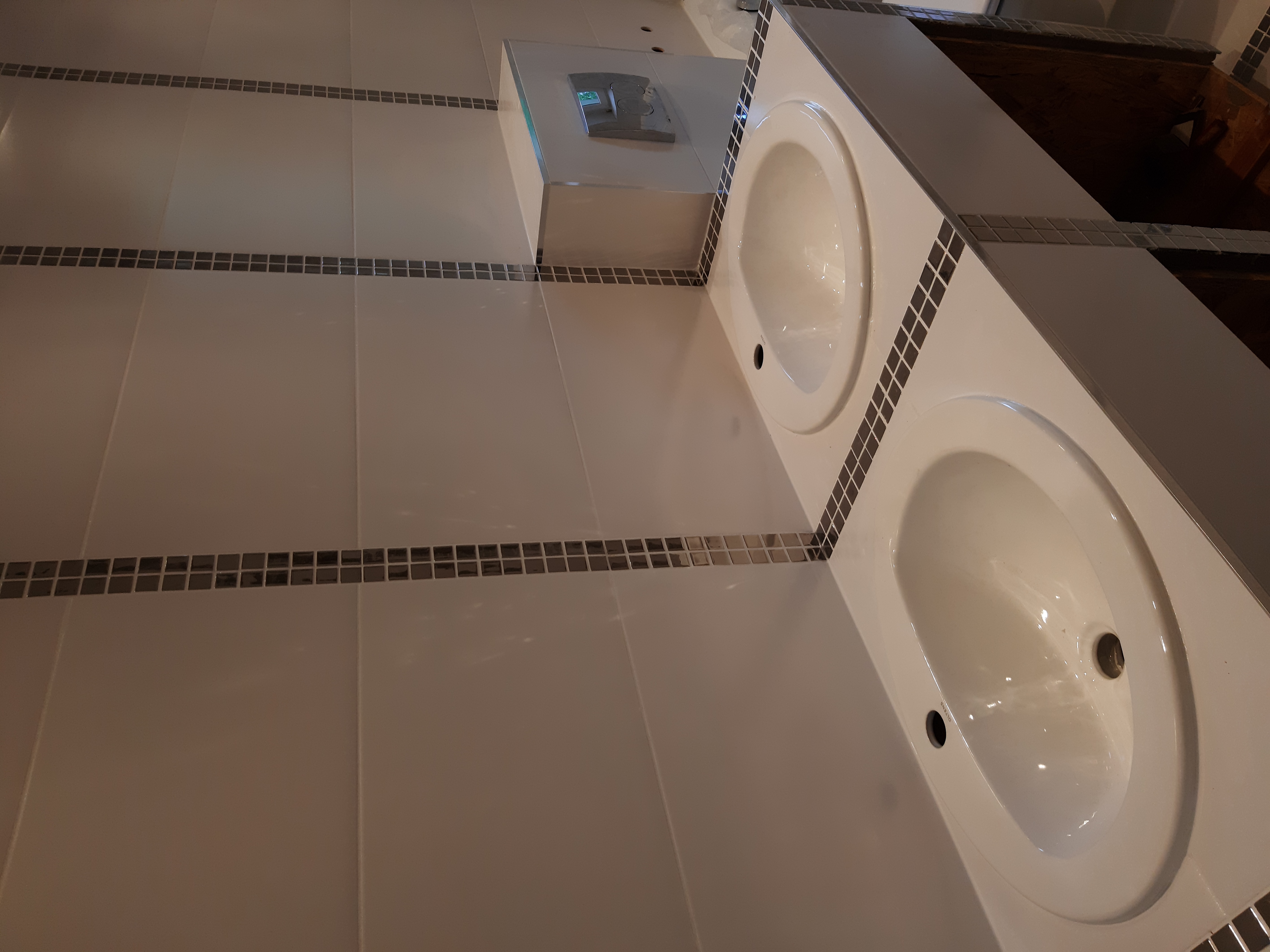 Dwie białe umywalki nablatowe w nowocześnie wykończonej łazience, z białymi płytkami i dekoracyjnymi pasami mozaiki.