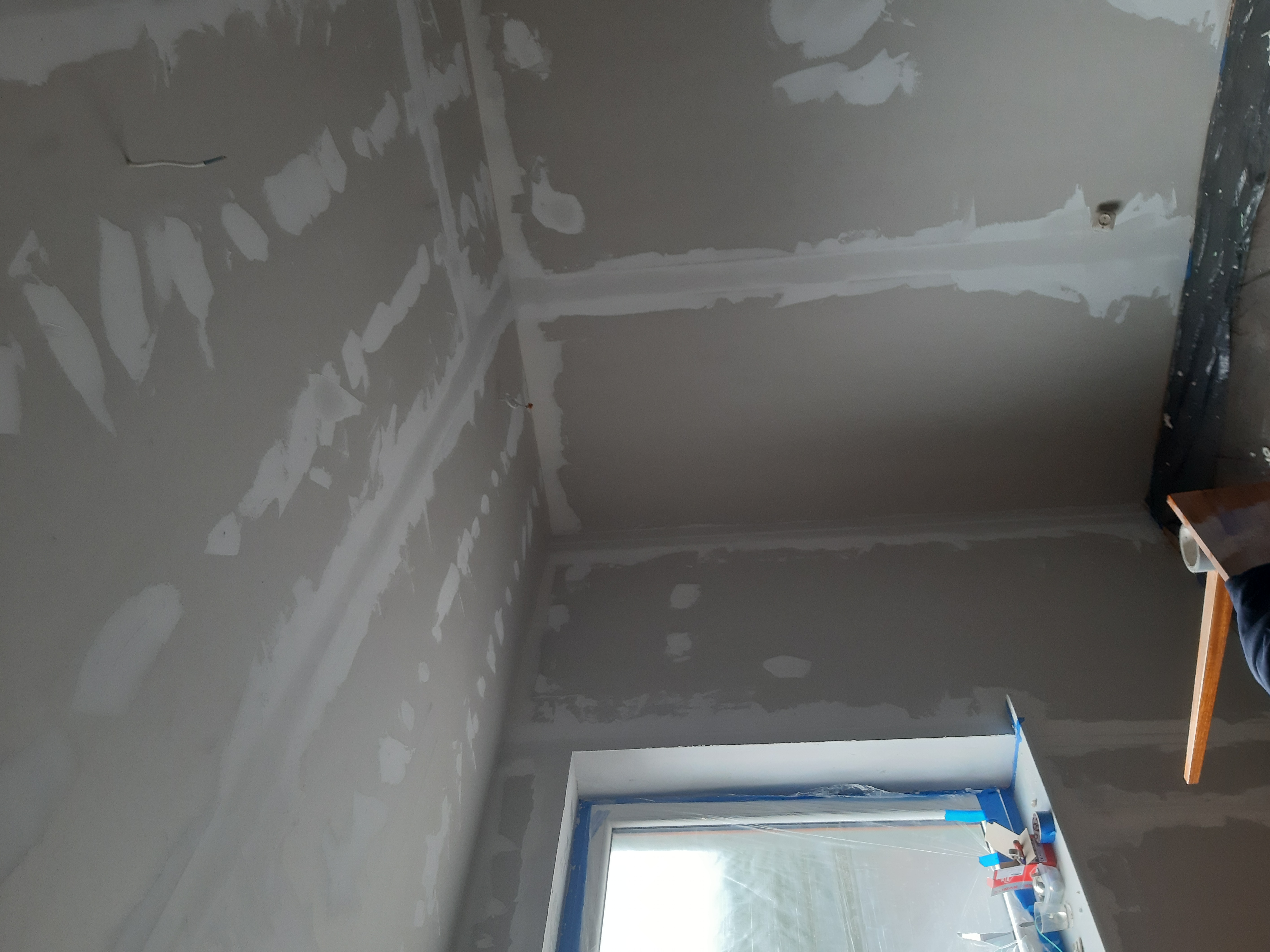 Szpachlowanie połączeń płyt karton-gips na suficie i ścianach, widoczne ślady po łatach, zabezpieczone okno folią i taśmą malarską, fragment drabiny w kadrze.