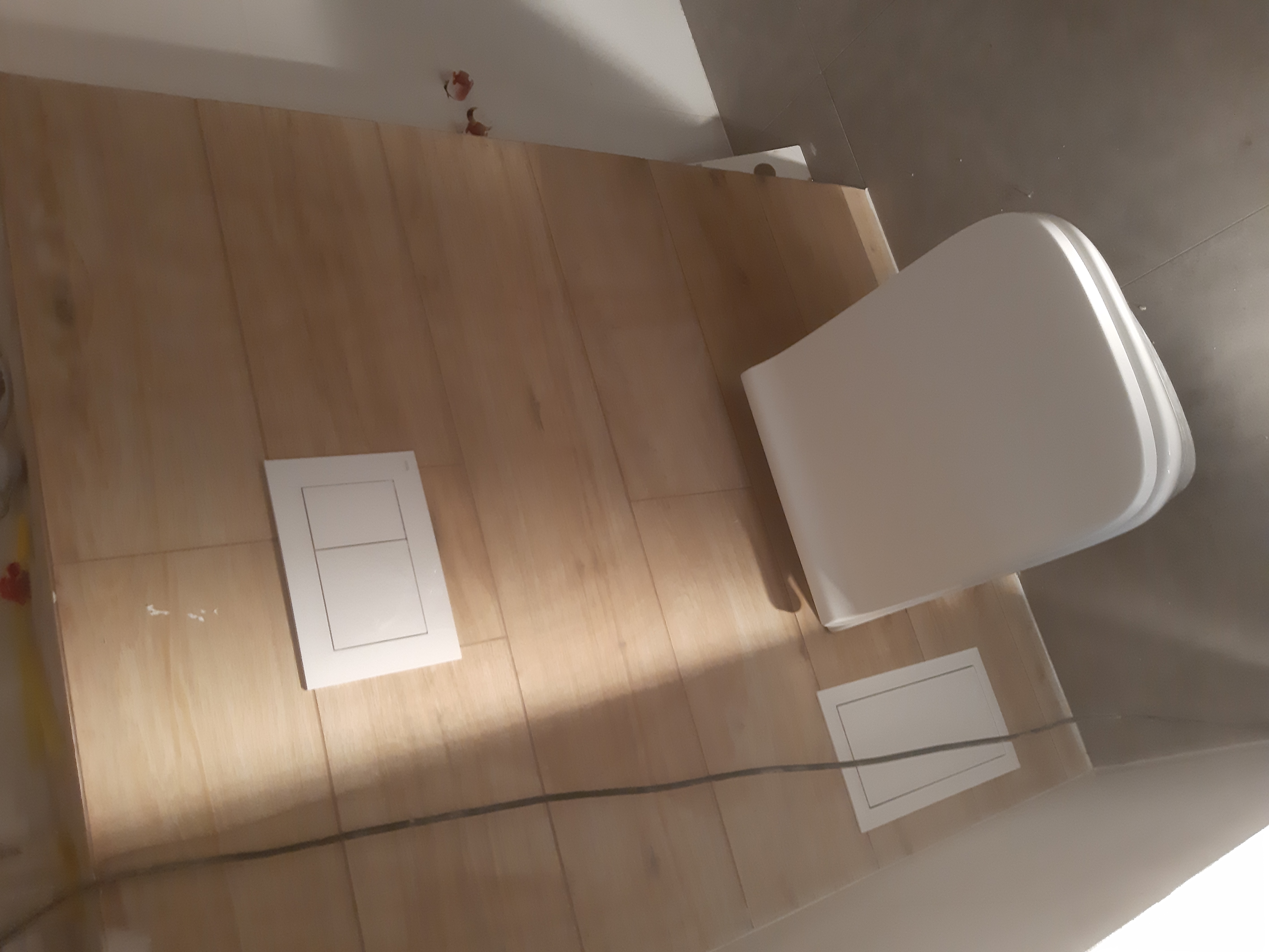 Ściana w łazience wyłożona płytkami imitującymi drewno, widoczny stelaż podtynkowy z przyciskiem spłukującym oraz fragment miski WC, podłączony czarny kabel elektryczny.