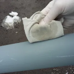 Ręka w lateksowej rękawicy czyści szmatką pomalowaną na szaro metalową rurę, widoczne drobne uszkodzenia farby i plamy na szmatce, w tle grudka topniejącego śniegu.