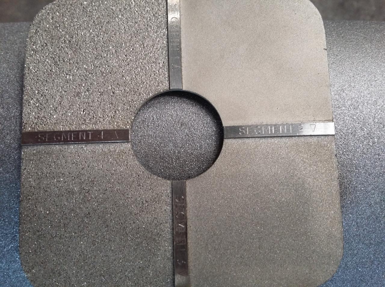 Metalowa płyta testowa z czterema segmentami o różnej fakturze powierzchni i oznaczeniami 'Segment 1 A', 'Segment 2 7', 'Segment 3 A', 'Segment 4 A', z okrągłym otworem pośrodku.
