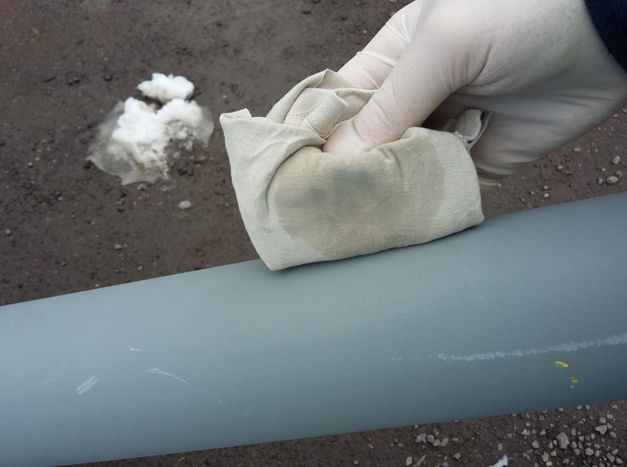 Ręka w lateksowej rękawicy czyści szmatką pomalowaną na szaro metalową rurę, widoczne drobne uszkodzenia farby i plamy na szmatce, w tle grudka topniejącego śniegu.