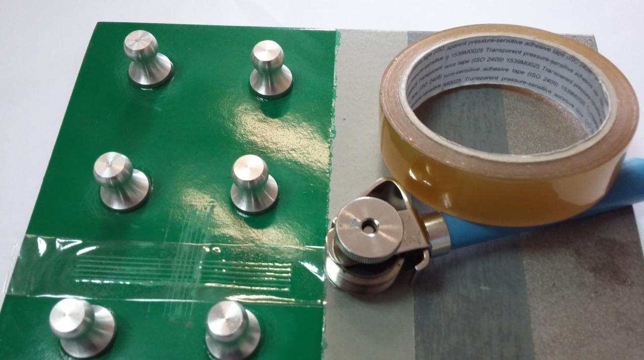 Test adhezji powłoki: metalowe trzpienie na zielonej powierzchni z nałożoną taśmą testową i narzędziem do dociskania taśmy.