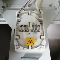 Splice tray z włóknami światłowodowymi ułożonymi wewnątrz, żółta naklejka ostrzegawcza przed promieniowaniem laserowym, widoczne kable w różnych kolorach.