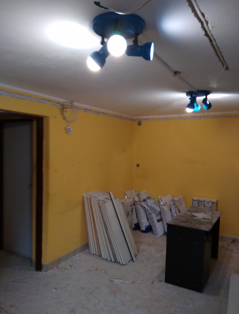 Żółty pokój w trakcie remontu, z niebieskimi reflektorami sufitowymi, workami z materiałami budowlanymi i stertą płyt przy ścianie.