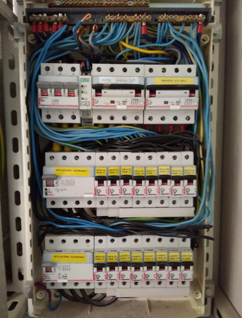 Rozdzielnica elektryczna z widocznymi bezpiecznikami, przewodami w kolorach niebieskim, czarnym i żółto-zielonym, oraz naklejkami identyfikacyjnymi na poszczególnych elementach, umieszczona...