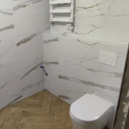 Nowoczesna toaleta wisząca w łazience wykończonej białymi płytkami imitującymi marmur z brązowymi żyłami, drewnopodobna podłoga ułożona w jodełkę, biały grzejnik drabinkowy.