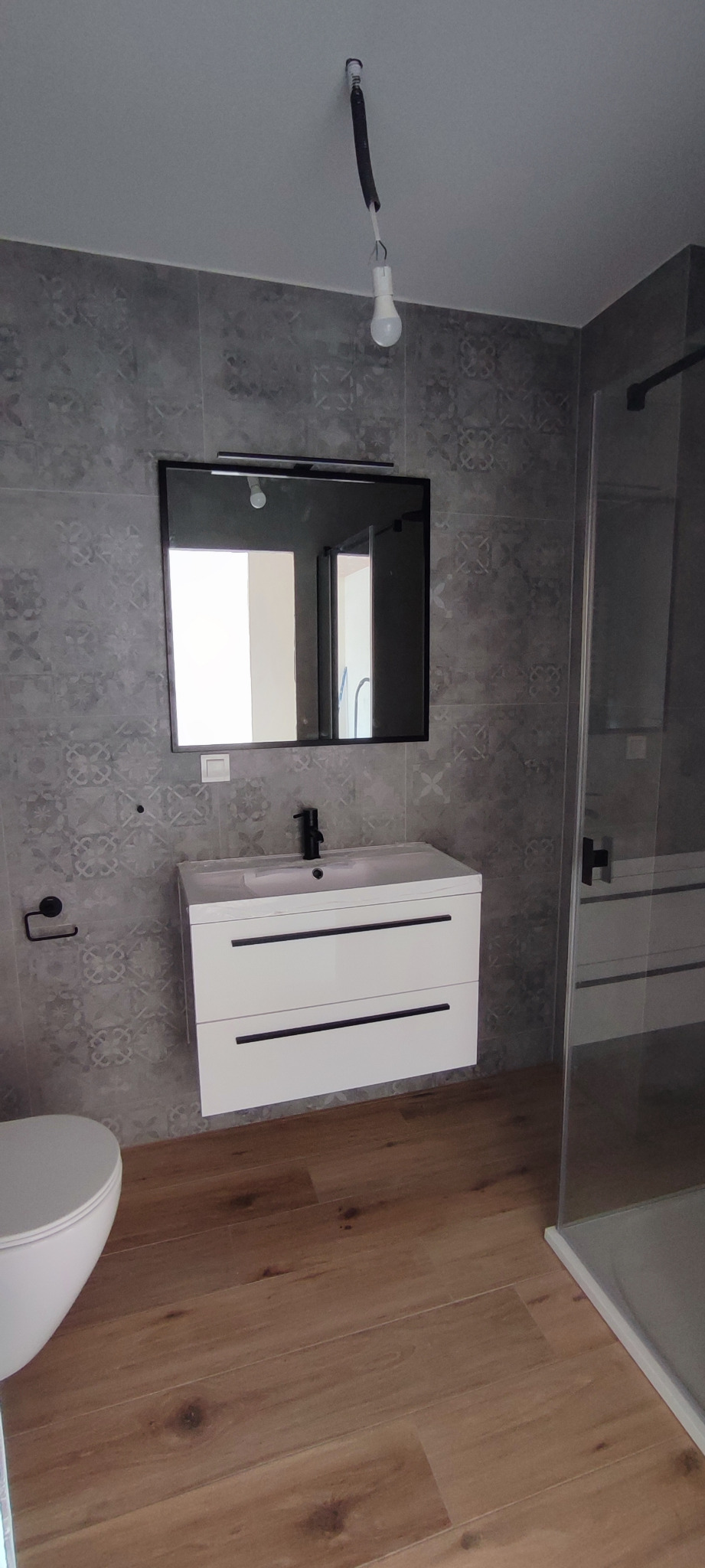 Wykończona łazienka z drewnopodobną podłogą, szarą mozaiką na ścianach, białą szafką pod umywalką z czarną armaturą oraz kabiną prysznicową z przezroczystego szkła. Na suficie wisi żarówka bez oprawy.