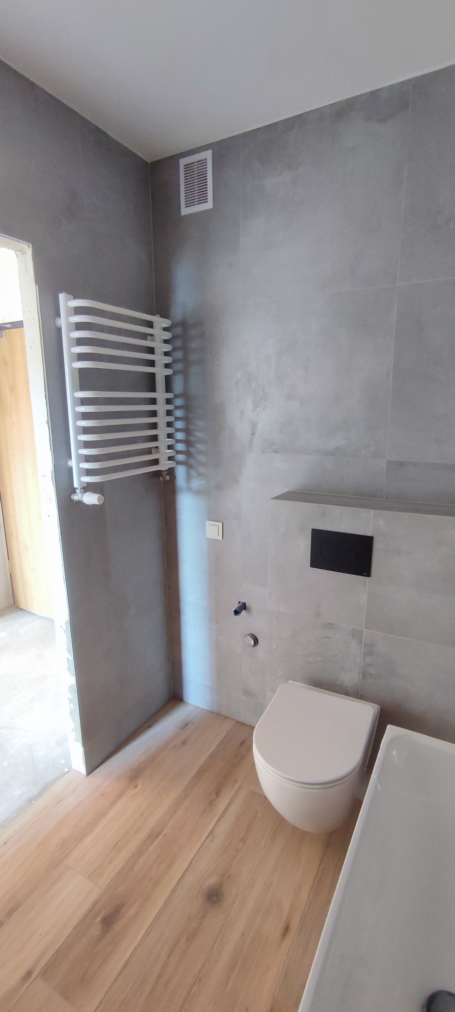 Wykończona łazienka w szarościach z drewnopodobną podłogą, widoczny grzejnik drabinkowy, toaleta wisząca i fragment brodzika.